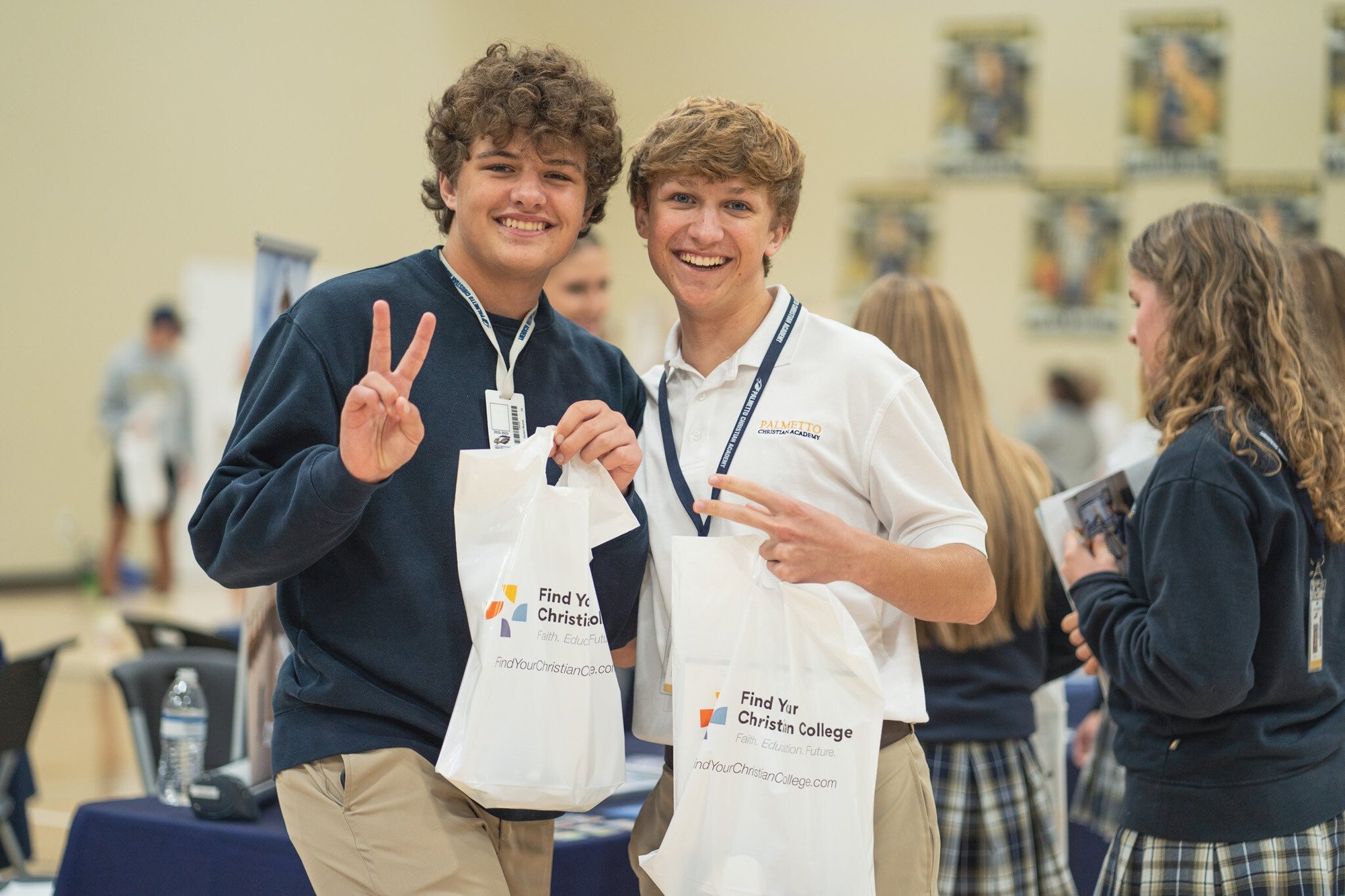 Two smiling students hold white bags and make peace signs at a college fair. Other attendees and booths are visible in the background.