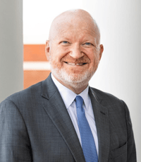 A bald man with a beard wearing a suit and blue tie smiles in front of a white column and brick wall background.