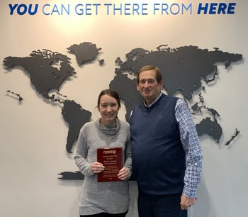 Two people stand in front of a world map mural. One holds a red plaque. The text above reads, "You can get there from here.