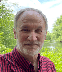 An older man with a beard and a checkered shirt smiles in front of a lush, green riverside background on a sunny day.