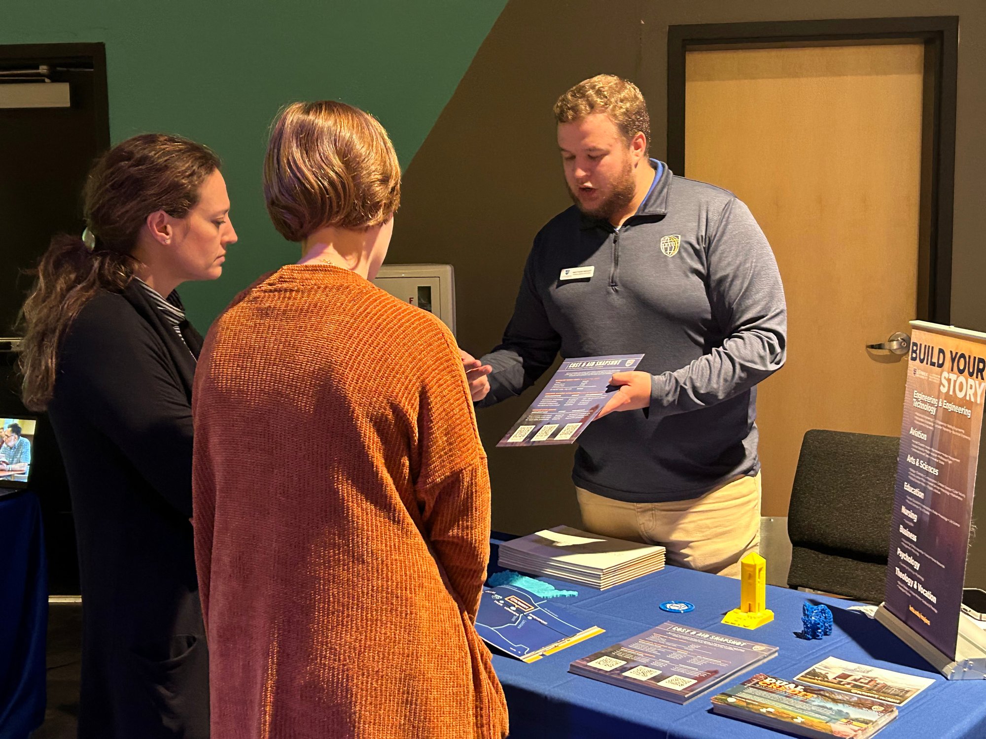 A man in a blue shirt is discussing materials on a table with two women at an indoor event.