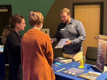A man in a blue shirt is discussing materials on a table with two women at an indoor event.
