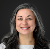 Smiling person with long gray hair, wearing a white top and hoop earrings, against a dark background.