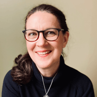 Woman with glasses and a ponytail, wearing a black sweater and a pendant necklace, smiling against a plain background.