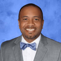 A person in a gray suit and blue bow tie against a blue background, smiling at the camera.