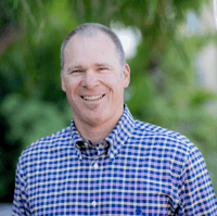 Smiling man wearing a blue and yellow checkered shirt stands outdoors with greenery in the background.